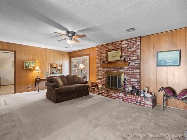 living room with ceiling fan, wooden walls, a textured ceiling, a brick fireplace, and light colored carpet