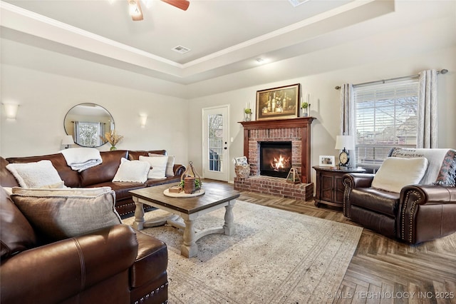 living room featuring parquet flooring, a brick fireplace, ornamental molding, a tray ceiling, and ceiling fan