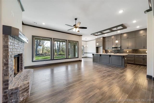 kitchen with dark wood-type flooring, backsplash, a fireplace, a center island with sink, and decorative light fixtures