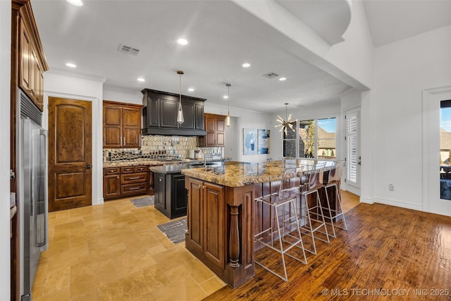 kitchen featuring a kitchen bar, high end fridge, tasteful backsplash, hanging light fixtures, and a kitchen island with sink