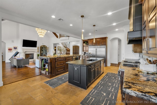 kitchen with built in fridge, decorative light fixtures, sink, exhaust hood, and a center island with sink