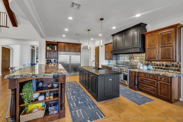 kitchen featuring an island with sink, sink, backsplash, premium appliances, and hanging light fixtures