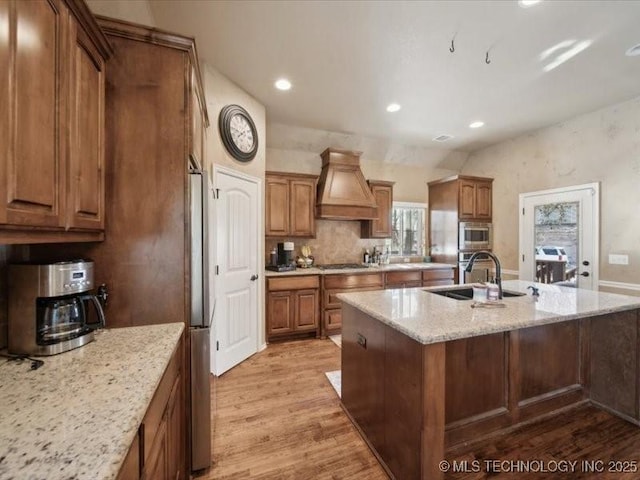 kitchen with sink, custom exhaust hood, stainless steel microwave, a kitchen island with sink, and light hardwood / wood-style floors