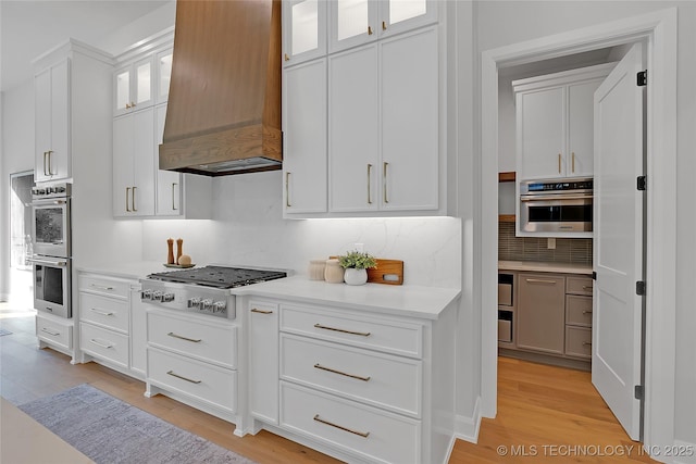 kitchen featuring light hardwood / wood-style flooring, custom range hood, white cabinets, stainless steel appliances, and backsplash