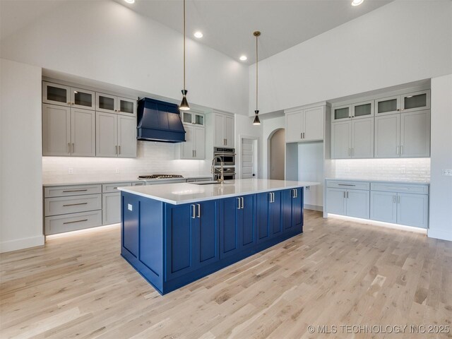 kitchen featuring appliances with stainless steel finishes, pendant lighting, an island with sink, custom exhaust hood, and light wood-type flooring