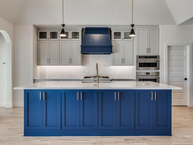 kitchen featuring arched walkways, custom range hood, a kitchen island with sink, vaulted ceiling, and light countertops