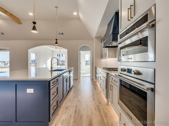 kitchen with sink, custom exhaust hood, hanging light fixtures, light hardwood / wood-style flooring, and appliances with stainless steel finishes