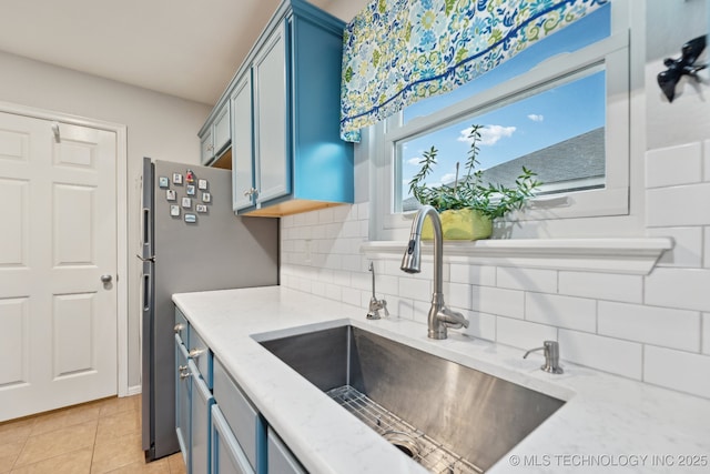 kitchen featuring sink, light tile patterned floors, blue cabinetry, stainless steel refrigerator, and decorative backsplash