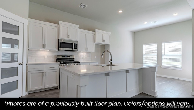 kitchen with sink, white cabinets, backsplash, stove, and a center island with sink