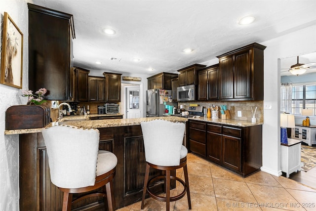 kitchen with dark brown cabinets, stainless steel appliances, tasteful backsplash, light stone counters, and a kitchen bar