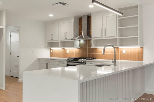 kitchen with wall chimney range hood, sink, white cabinetry, gas range, and kitchen peninsula