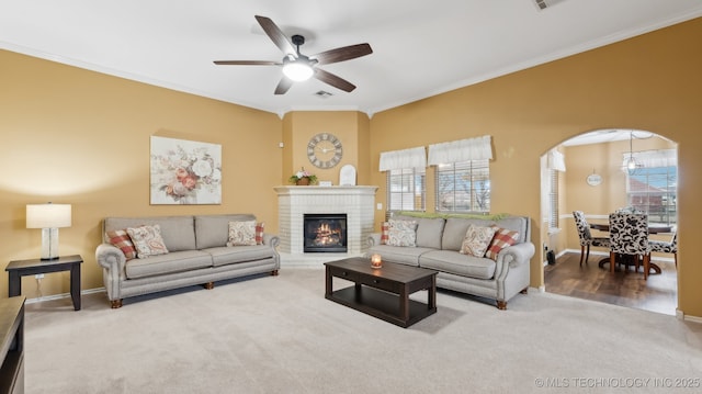 living room featuring crown molding, carpet flooring, ceiling fan with notable chandelier, and a brick fireplace