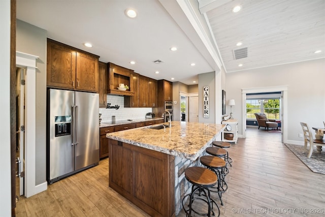 kitchen with high end fridge, sink, tasteful backsplash, light stone countertops, and a kitchen island with sink