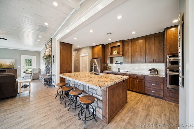 kitchen featuring a breakfast bar, an island with sink, backsplash, stainless steel appliances, and light stone countertops