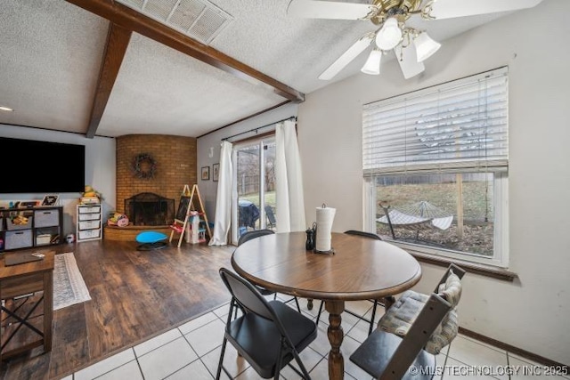 dining room with ceiling fan, a fireplace, a textured ceiling, light tile patterned flooring, and beamed ceiling