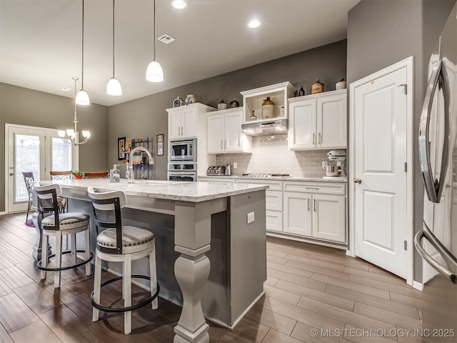 kitchen with white cabinetry, decorative light fixtures, stainless steel appliances, and an island with sink