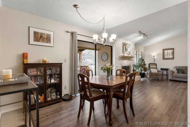 dining area featuring lofted ceiling, wood-type flooring, a chandelier, a brick fireplace, and a textured ceiling