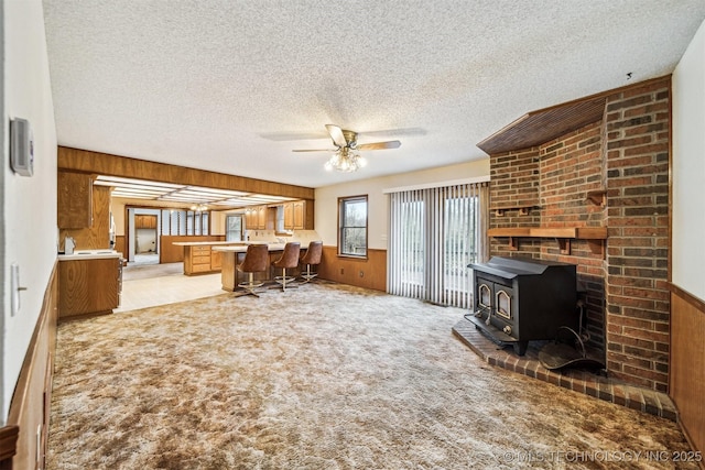 unfurnished living room featuring wood walls, light colored carpet, a textured ceiling, a wood stove, and ceiling fan