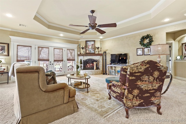 living room featuring french doors, a stone fireplace, crown molding, a raised ceiling, and light colored carpet