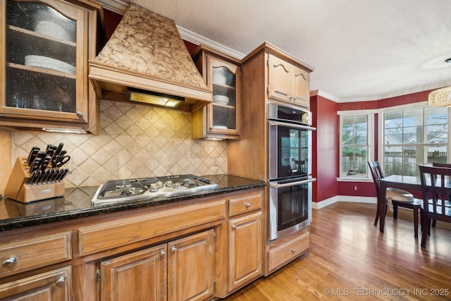 kitchen featuring appliances with stainless steel finishes, backsplash, light hardwood / wood-style floors, custom exhaust hood, and dark stone counters