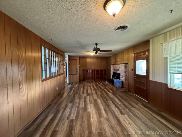 unfurnished living room with a fireplace, wood walls, ceiling fan, dark wood-type flooring, and a textured ceiling