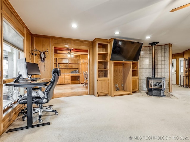 carpeted home office with ceiling fan, a wood stove, and wooden walls