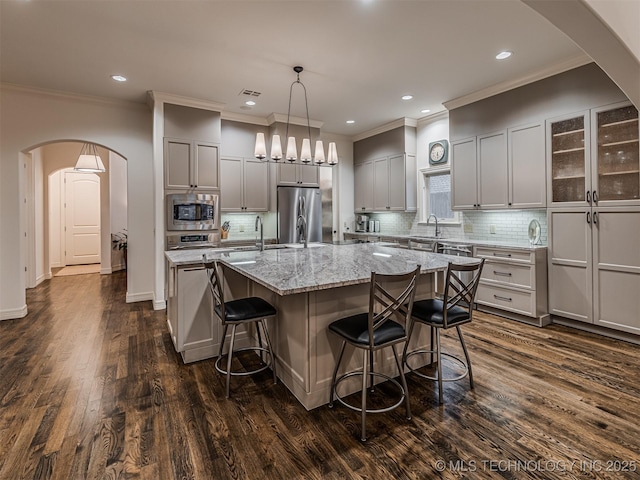 kitchen with appliances with stainless steel finishes, light stone counters, dark hardwood / wood-style flooring, hanging light fixtures, and a kitchen island with sink