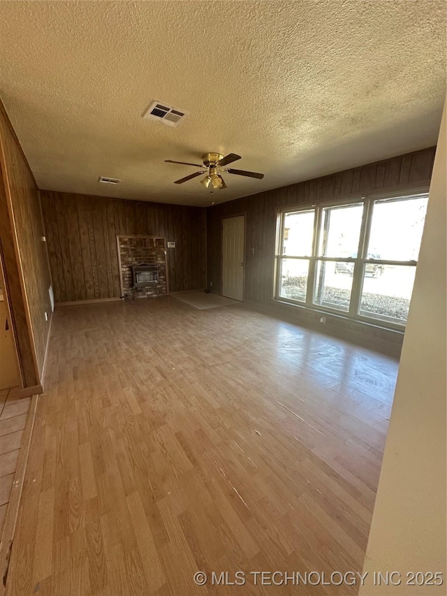 unfurnished living room with light hardwood / wood-style flooring, ceiling fan, a textured ceiling, a brick fireplace, and wood walls