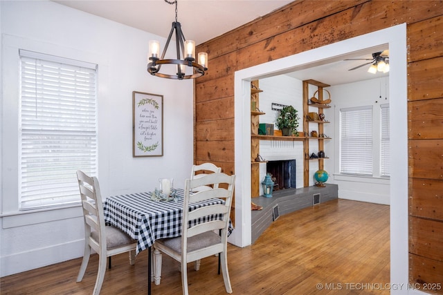 dining room with wood-type flooring, ceiling fan with notable chandelier, a fireplace, and wood walls