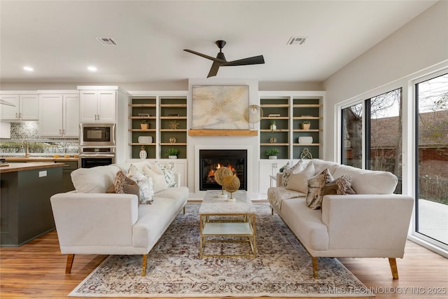 living room featuring visible vents, a ceiling fan, a lit fireplace, and light wood finished floors