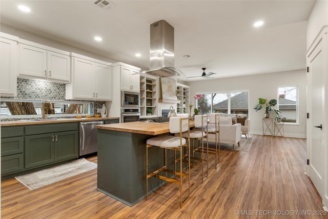 kitchen with green cabinetry, a sink, island range hood, appliances with stainless steel finishes, and butcher block counters