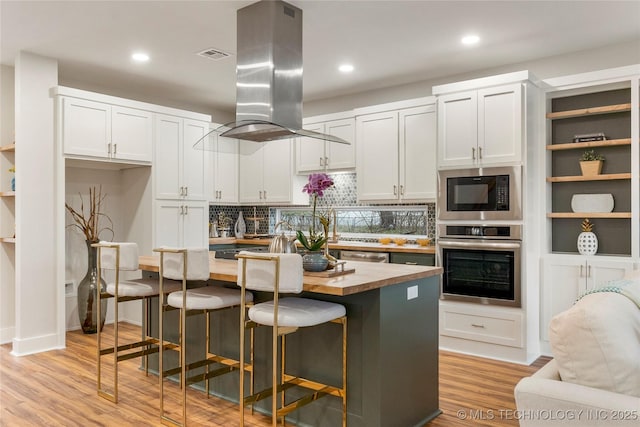 kitchen with stainless steel appliances, light wood-style floors, island exhaust hood, tasteful backsplash, and wooden counters