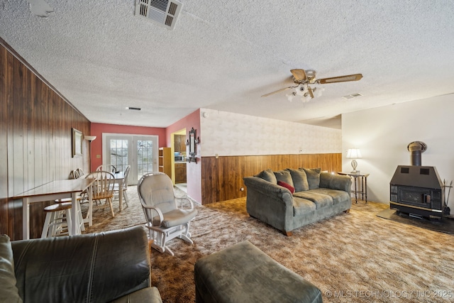 carpeted living room featuring ceiling fan, wood walls, a textured ceiling, and a wood stove