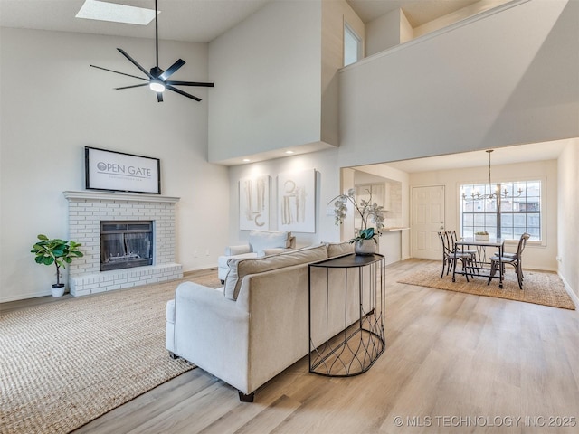 living room with a skylight, baseboards, light wood-style floors, a brick fireplace, and ceiling fan with notable chandelier