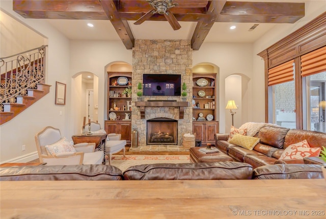 living room with built in shelves, ceiling fan, a stone fireplace, and beamed ceiling