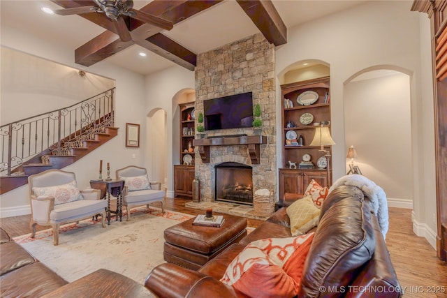 living room with ceiling fan, beam ceiling, a stone fireplace, and light hardwood / wood-style flooring