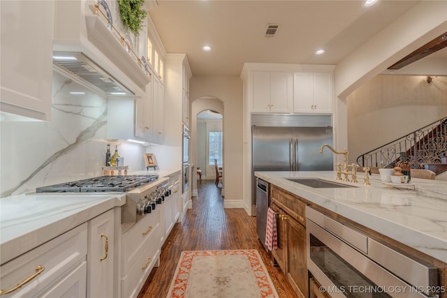 kitchen featuring wall chimney range hood, built in appliances, sink, and white cabinets