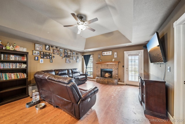 living room featuring a textured ceiling, a brick fireplace, light wood-type flooring, a tray ceiling, and ceiling fan