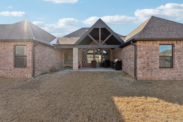 back of property with a yard, brick siding, a patio area, and a shingled roof