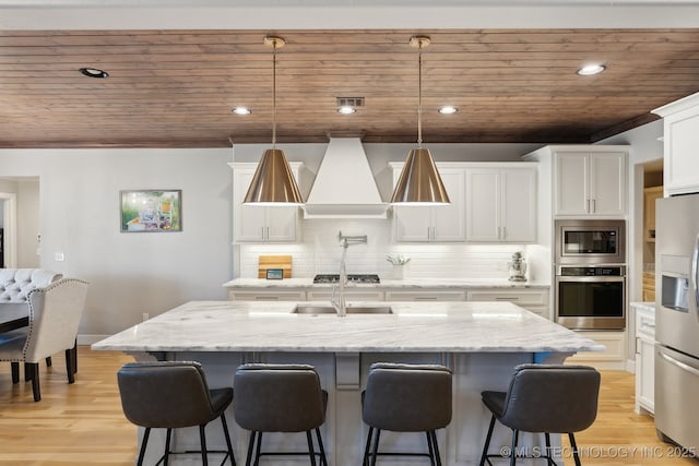 kitchen featuring white cabinets, a kitchen island with sink, stainless steel appliances, premium range hood, and pendant lighting