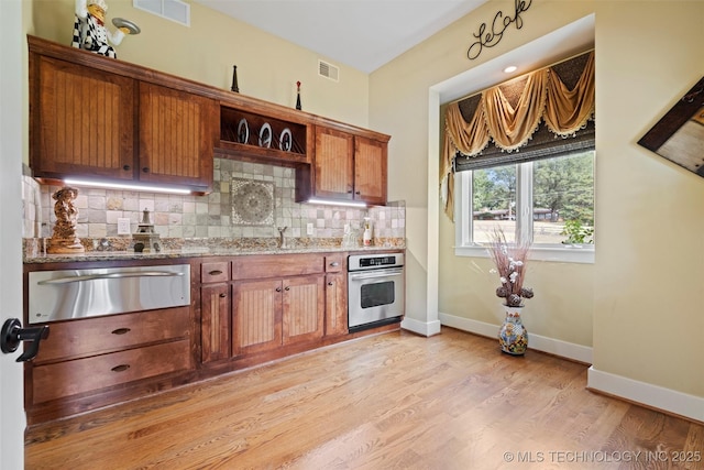 kitchen with tasteful backsplash, light stone countertops, stainless steel oven, and light wood-type flooring