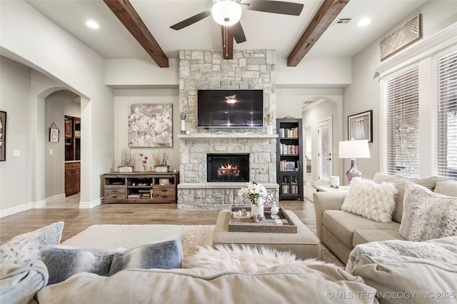 living room featuring beamed ceiling, ceiling fan, a stone fireplace, and light wood-type flooring