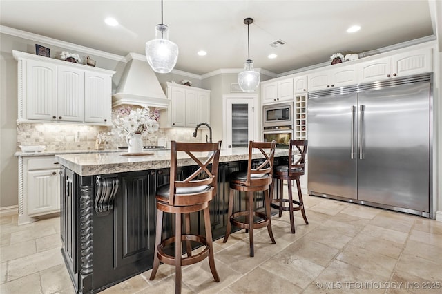 kitchen with custom exhaust hood, built in appliances, hanging light fixtures, a center island with sink, and white cabinets