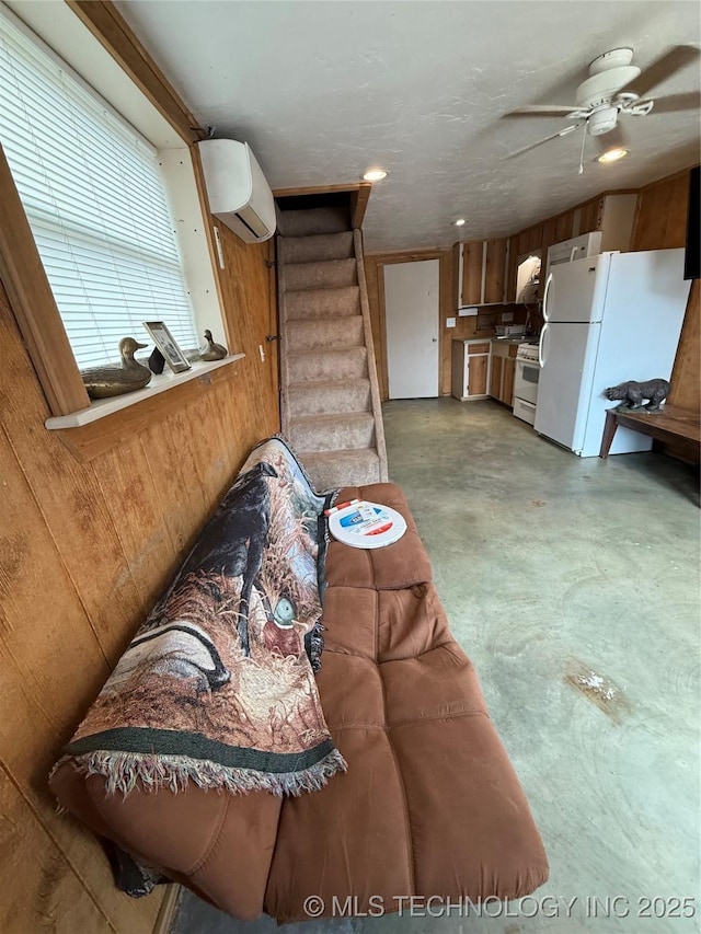 living room with a wall unit AC, stairway, wooden walls, a ceiling fan, and concrete floors