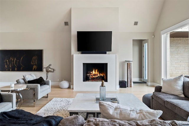 living area with a warm lit fireplace, light wood-style flooring, visible vents, baseboards, and vaulted ceiling