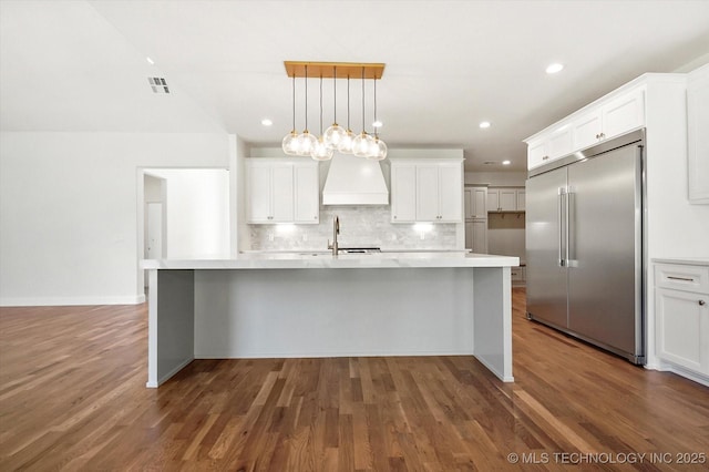 kitchen featuring pendant lighting, built in refrigerator, custom exhaust hood, and a center island with sink