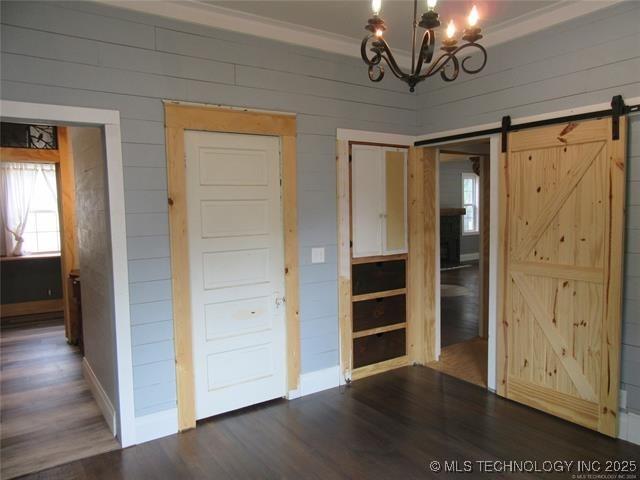 unfurnished bedroom with wood walls, ornamental molding, dark hardwood / wood-style flooring, a notable chandelier, and a barn door