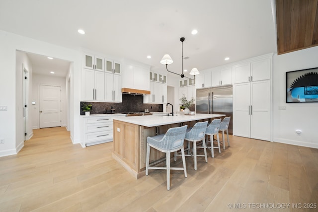 kitchen with backsplash, built in fridge, white cabinets, hanging light fixtures, and an island with sink