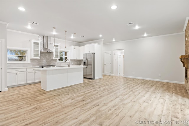 kitchen featuring stainless steel refrigerator with ice dispenser, white cabinetry, pendant lighting, a kitchen island with sink, and wall chimney range hood