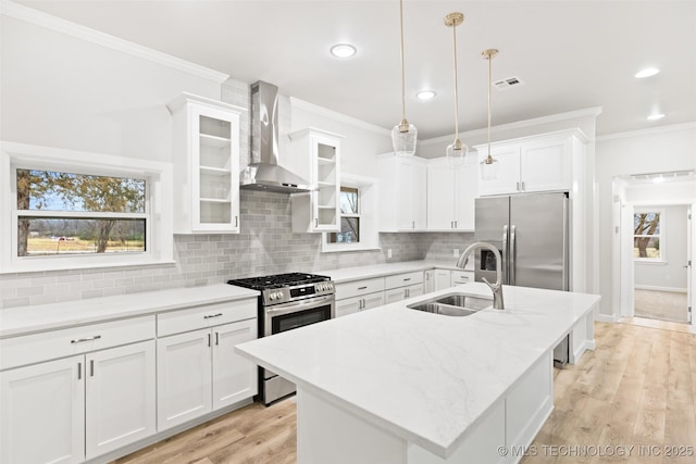 kitchen featuring wall chimney range hood, sink, appliances with stainless steel finishes, hanging light fixtures, and an island with sink